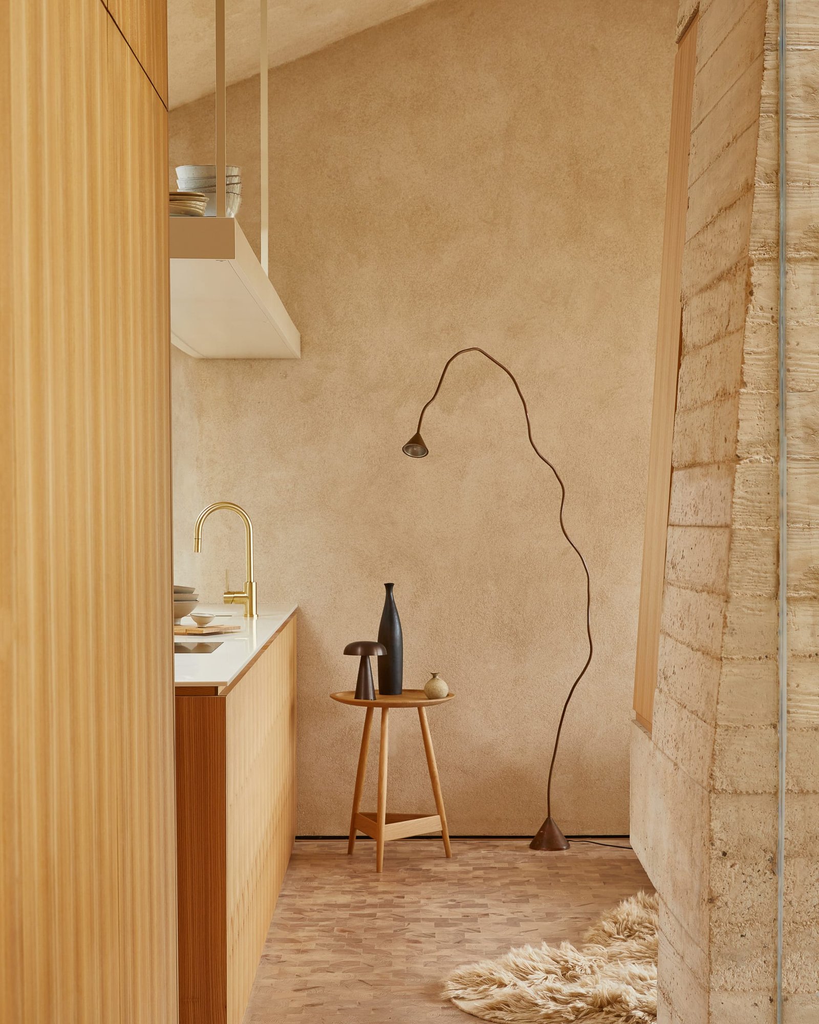 Clay-plastered wall at The Makers Barn with a slender floor lamp, beside a fluted timber run and shuttered concrete.