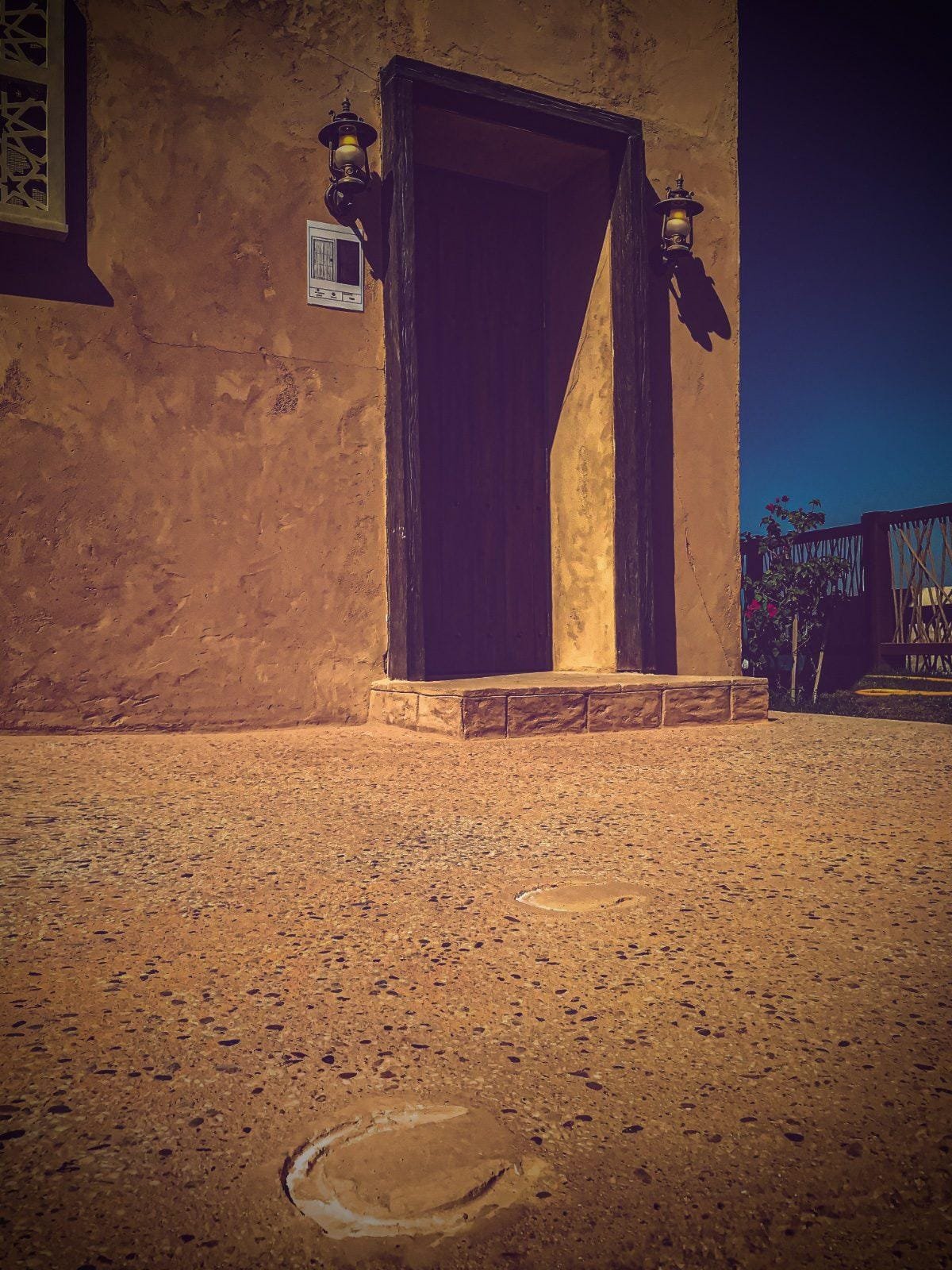 Ochre exposed aggregate court with imprinted horseshoe prints, beside an earthen-plastered building