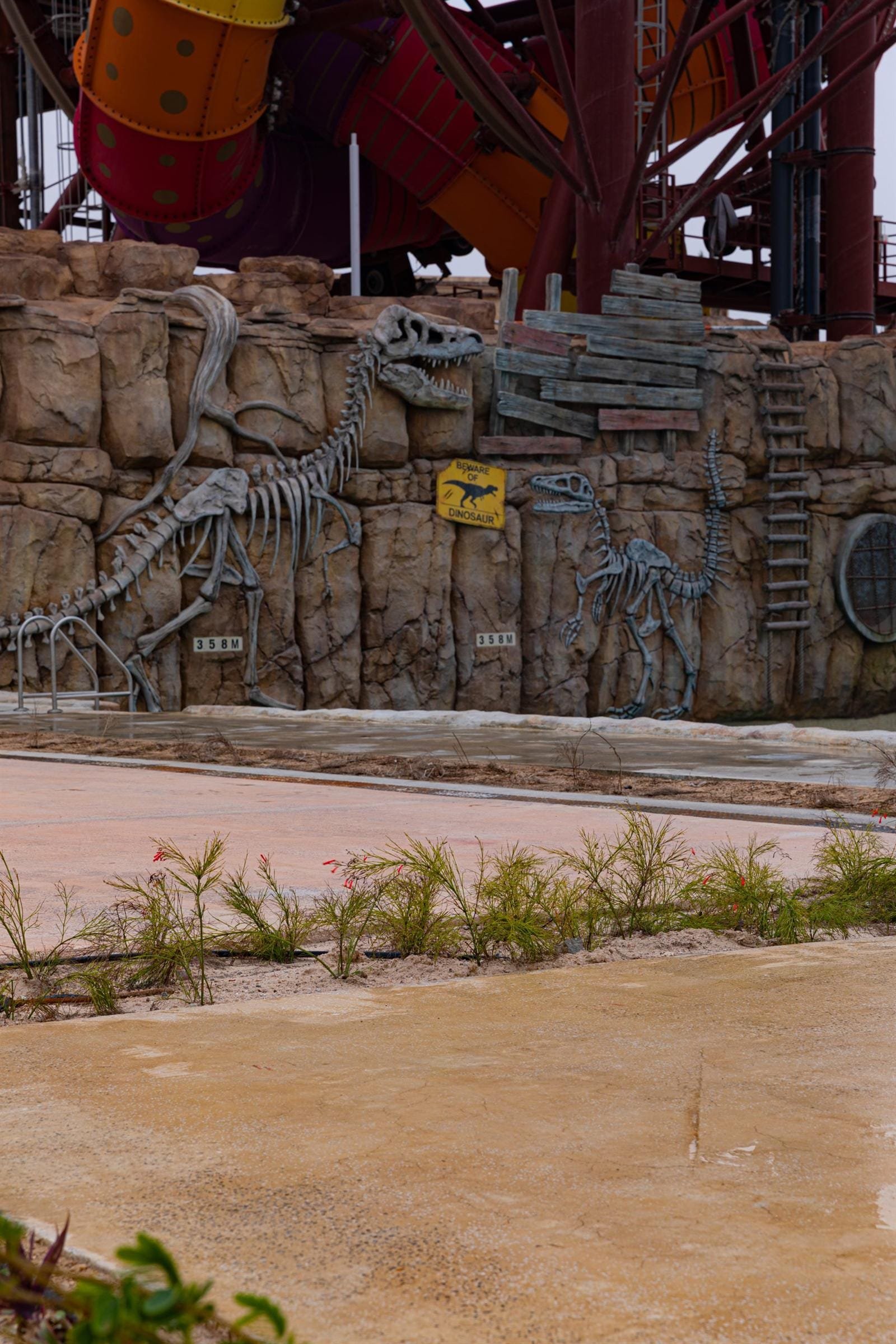 Two-tone pink and ochre exposed aggregate paths beside a rockwork wall with sculpted dinosaur skeletons at Meryal Waterpark