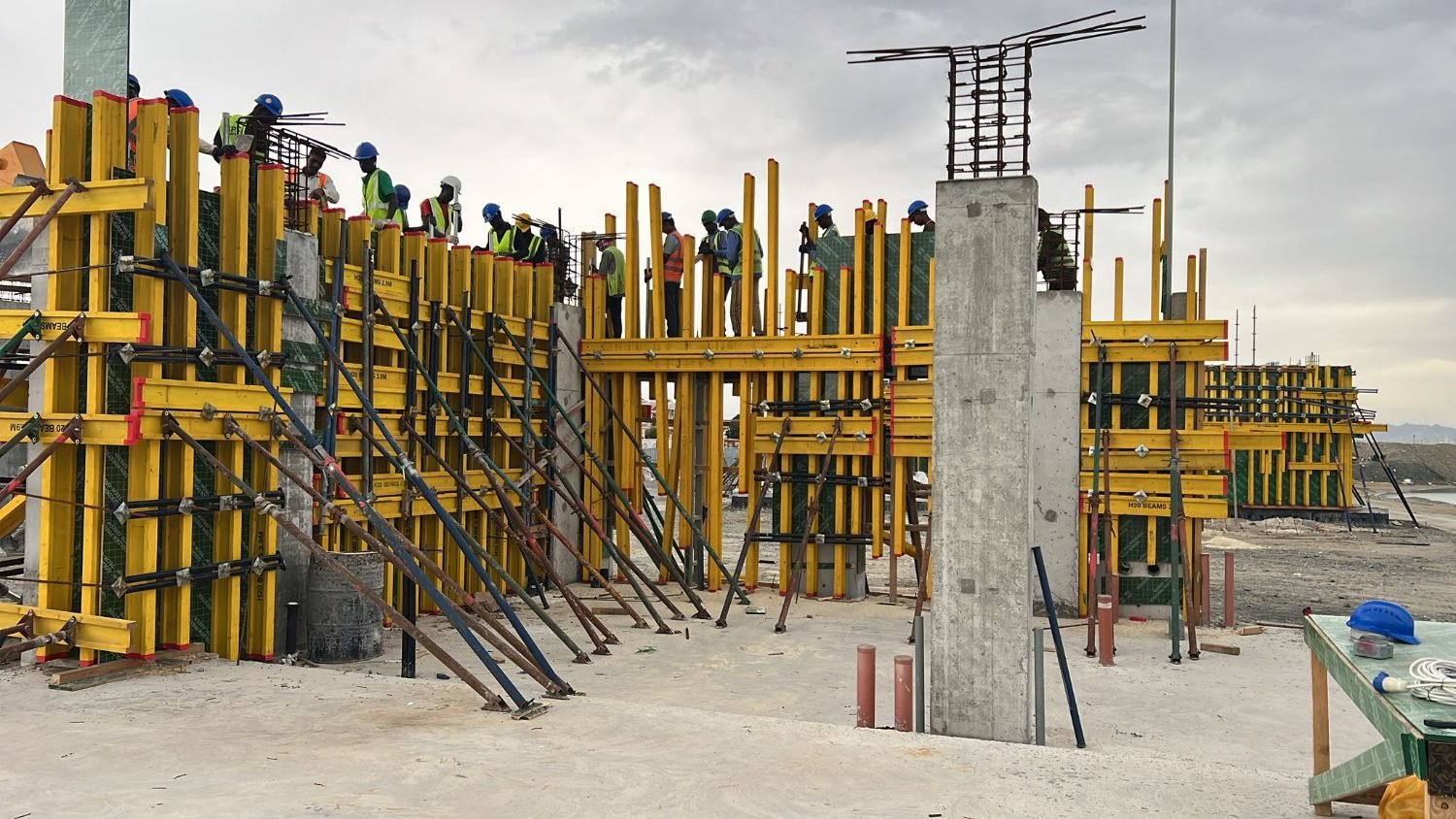 Crews in hi-vis on yellow H20 beam formwork at Kalba Beach with the Gulf of Oman behind them under a heavy grey sky.