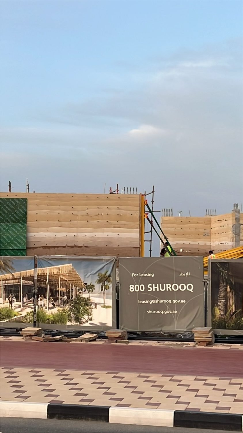 Rammed earth walls rising behind Shurooq site hoarding at Kalba Beach, the tamped bands visible above the leasing boards.
