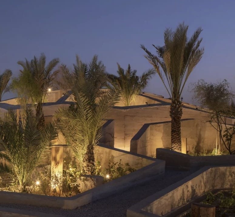 In-situ rammed earth garden walls uplit at dusk, date palms rising between them at the UAE Pavilion