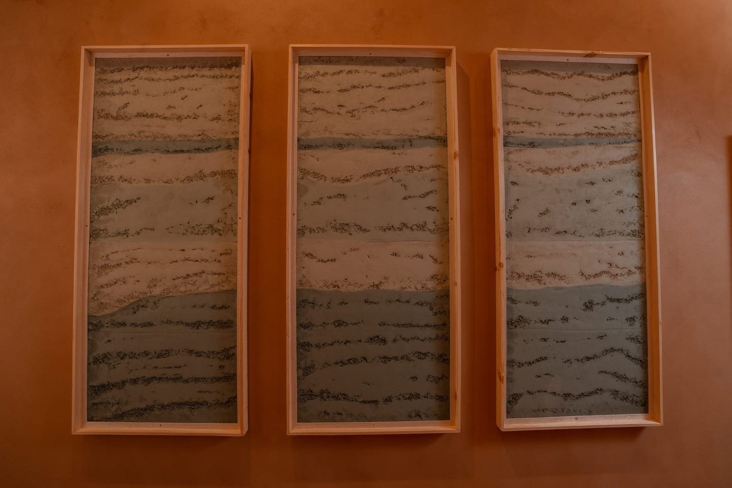Three framed rammed earth plaster panels at the Conmarble showroom, cream and grey-green tamped bands with open grain.